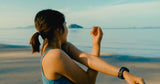 oung Asian woman in sportswear stretching by the sea, enjoying the warm morning sun, preparing for workout on serene beach