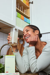 happy customer drinking Modern Dose Strawberry Biotic Blend in the kitchen with Modern Dose supplement box and stick packs on the table - www.moderndose.com