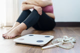 Overweight woman sitting on the floor upset with a scale and measuring tape next to her
