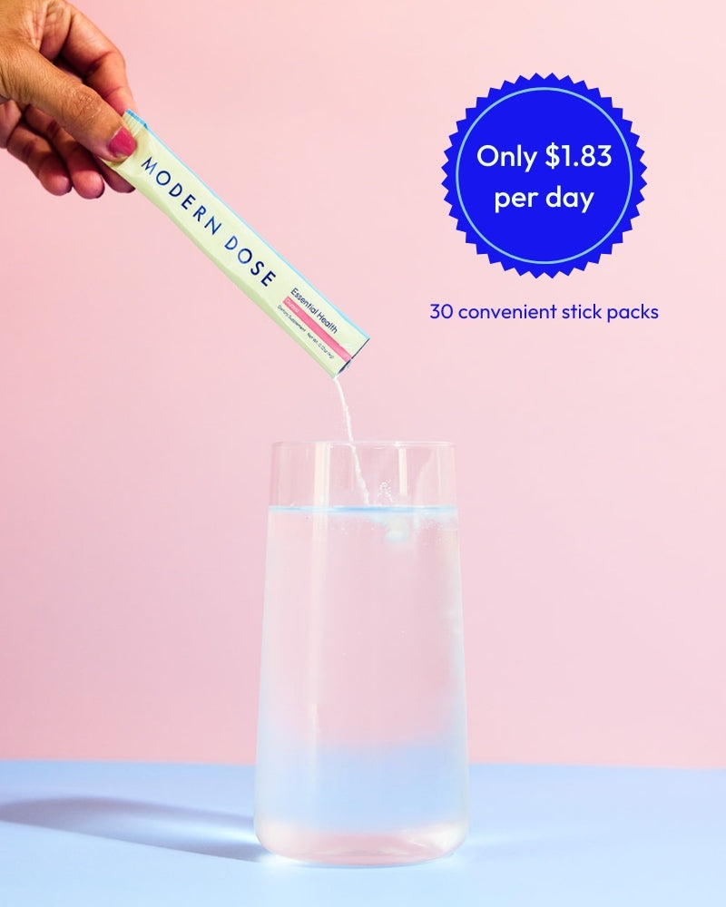 Hand pouring Modern Dose Essential Health into a glass of water with promotional text stating its only $1.83 per day, with a pink background - www.moderndose.com